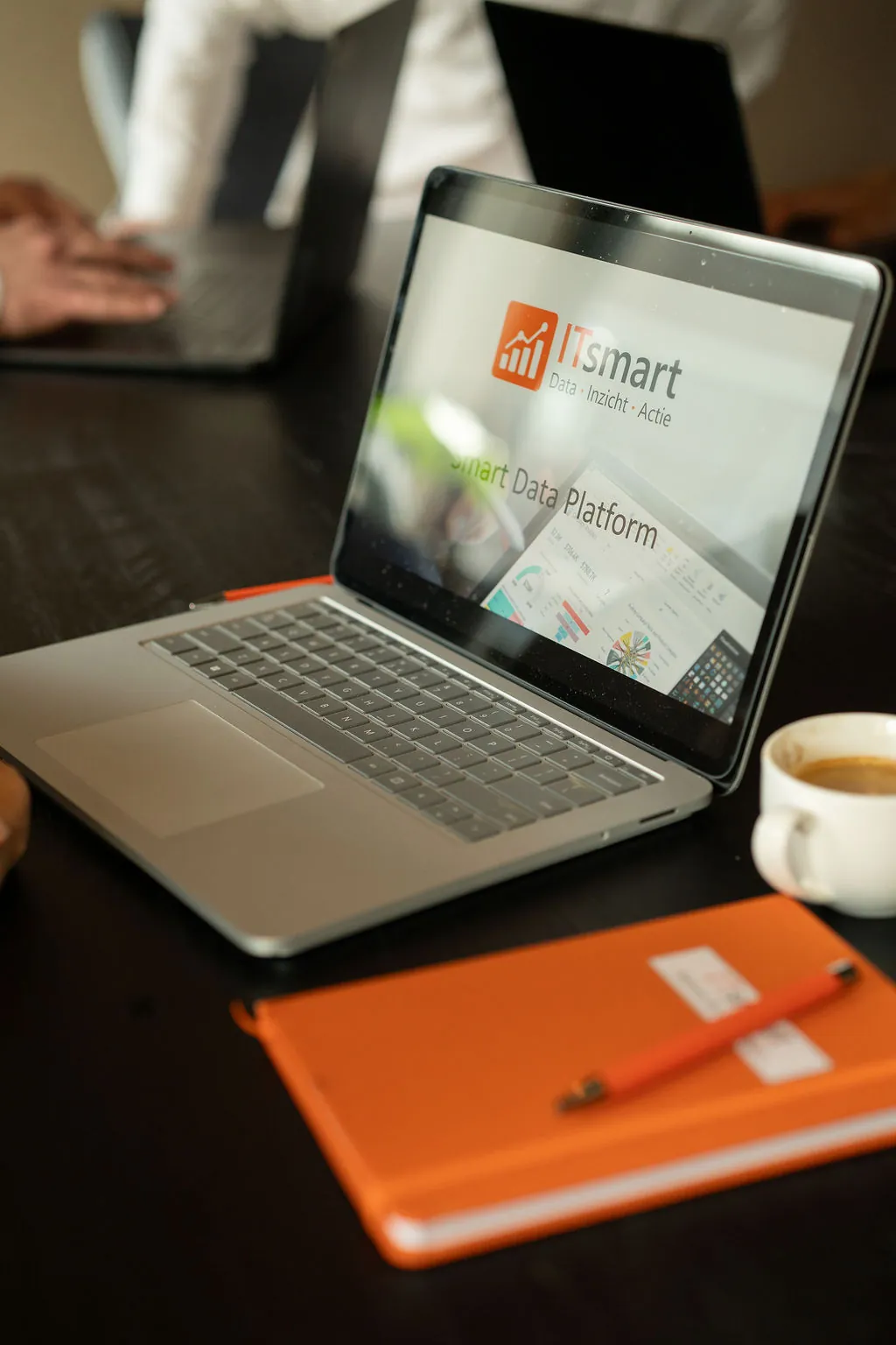 Close-up van een laptop met het ITsmart Smart Data Platform en een dashboard, naast een oranje notitieboek en koffie